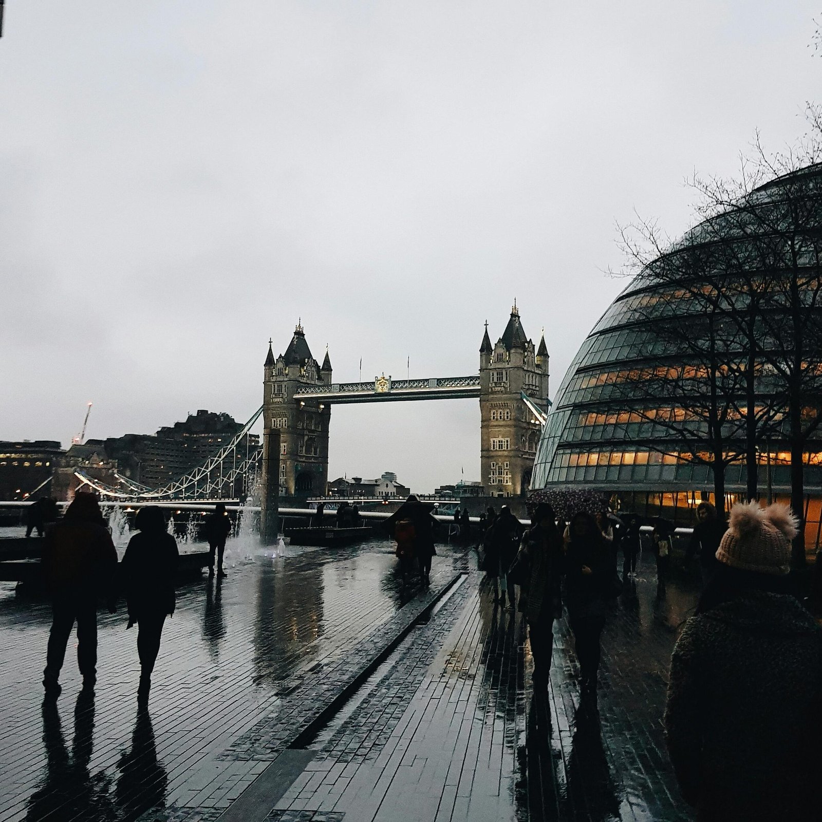 Rainy view of London's Tower Bridge and City Hall with pedestrians on a wet day.